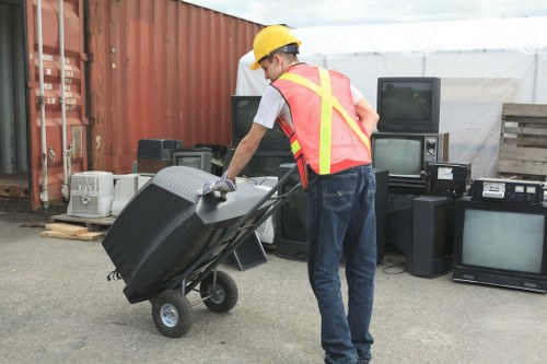 A worker wearing a yellow safety helmet, white T-shirt, and a high-visibility vest is pushing a black wheeled rubbish bin on a grey concrete surface. Behind him, there are various discarded electronic appliances, including old TVs, stereo systems, and other electronic waste, stacked against a red container and a wooden pallet. The scene is outdoors, with a white tent structure partially visible in the background, suggesting a waste collection or clearance site in a London area such as Mayfair. The worker appears to be preparing to load the rubbish for removal, illustrating professional waste management services offered by Office Clearance Mayfair for commercial or office waste disposal involving electronic items and general refuse. The environment is well-lit with natural daytime light, and the overall setting emphasizes the process of clearing and managing waste for city-based clients.