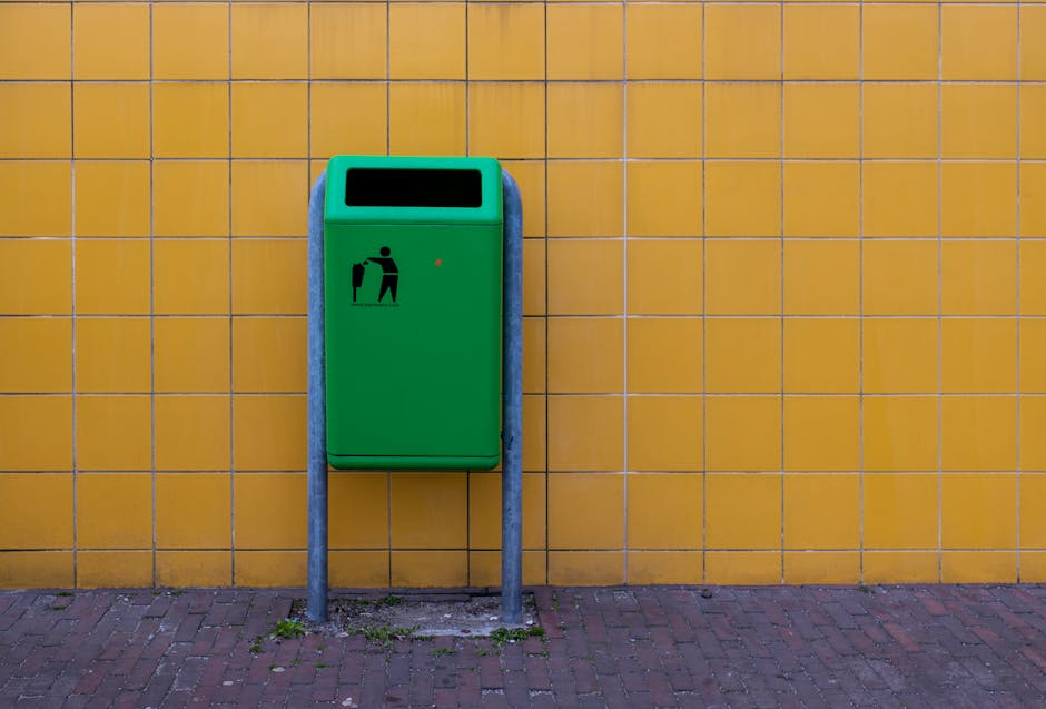 A green public waste bin mounted on two metal posts standing against a vibrant mustard-yellow tiled wall. The bin is rectangular with a wide, open slot at the top for depositing waste, and it features