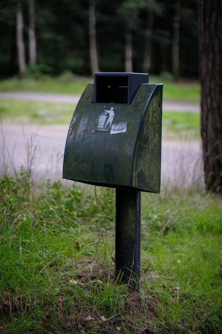 A green public waste bin mounted on two metal posts standing against a vibrant mustard-yellow tiled wall. The bin is rectangular with a wide, open slot at the top for depositing waste, and it features